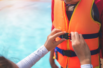 Asian mother helping her son to wear life jacket before playing in swimming pool, Selective focus.